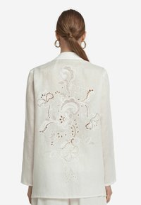 Vit linnet blazer med långa ärmar, med intrikata blommönster broderi och utskärningar på baksidan. Slät textur.
