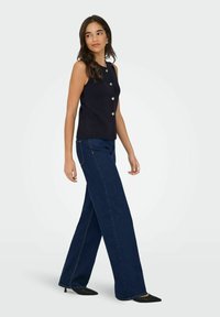 Marinefarbene ärmellose Bluse mit metallischen Herzknöpfen, kombiniert mit weit geschnittenen, dunkelblauen Jeans mit dezentem Stichmuster und spitzen schwarzen Schuhen.