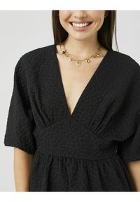 Robe noire texturée avec un profond décolleté en V, manches courtes bouffantes et taille froncée. Détails remarquables : des ornements subtils et un collier superposé.