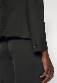 Persona con chaqueta y pantalones de traje negro a medida, de pie contra un fondo claro y sencillo, con la mano relajada al costado.