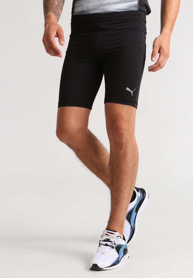 Homme portant un short de compression Puma noir et des chaussures de sport blanches avec des accents noirs et bleus, debout avec une jambe croisée.