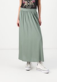 Maxi gonna verde chiaro realizzata in tessuto morbido con vita elastica, abbinata a sneakers bianche alte. Design semplice e fluido senza motivi.