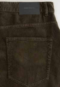 Bruin corduroy stof met verticale ribbels, een gestikte zak en een zwart label met de tekst "DENIM SUPPLY Co." op de tailleband.