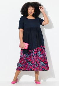 Marineblaues Kleid mit kurzen Ärmeln und quadratischem Ausschnitt; geraffte Taille mit Blumenmuster in Pink und Blau, kombiniert mit einer pinken Clutch und Schuhen.