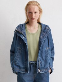 Denimjacke mit Kapuze, Reißverschluss, zwei vorderen Taschen und Kontraststichen; kombiniert mit einem gerippten grünen Oberteil.