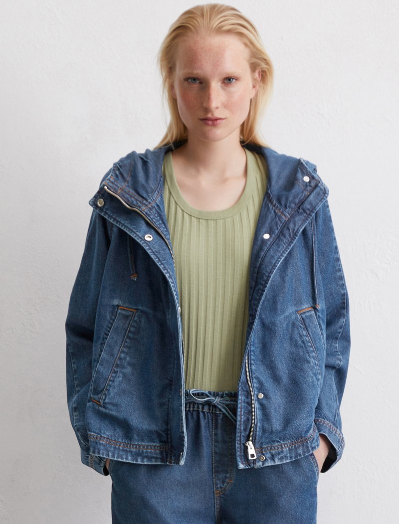 Denimjacke mit Kapuze, Reißverschluss, zwei vorderen Taschen und Kontraststichen; kombiniert mit einem gerippten grünen Oberteil.