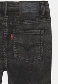 Pantalones vaqueros negros de mezclilla con un acabado texturizado. Incluyen un bolsillo trasero, una etiqueta roja y un parche de cuero negro con detalles de la marca.