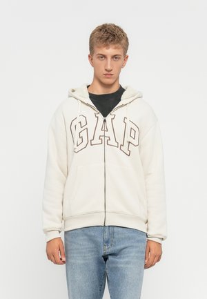 Jeune homme portant un sweat à capuche zippé beige clair de la marque GAP avec une capuche en polaire et un jean bleu clair, se tenant devant un fond blanc uni.