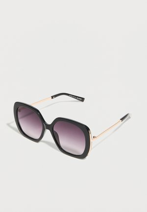 Übergroße quadratische Sonnenbrille mit schwarzem Rahmen, verlaufenden dunkelgrauen Gläsern und schlanken, goldfarbenen Metallbügeln auf weißer Oberfläche.