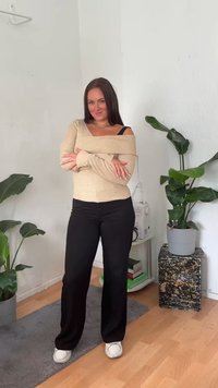 Maglione beige a spalle scoperte con coste, pantaloni neri a gamba larga e sneakers bianche. Piante verdi e un tavolo in pietra decorato sullo sfondo.