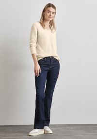 Cremefarbener V-Ausschnitt-Pullover aus strukturiertem Stoff, kombiniert mit dunkelblauen Schlagjeans und weißen Sneakern. Einfaches Design ohne auffällige Muster.