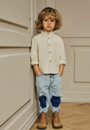 Beige skjorte med lange ærmer, blå denim jeans med mørke pletter, og brune ankelstøvler. Stående mod en struktureret væg.
