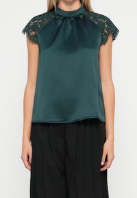 Femme portant un chemisier sans manches en satin vert foncé avec des manches courtes en dentelle et un col froncé, associé à un pantalon noir à jambes larges.