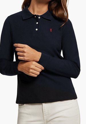 Kobieta ubrana w granatową koszulkę polo z długim rękawem z czerwonym wyszywanym logo i kremowe spodnie, z rękami skrzyżowanymi na wysokości talii.