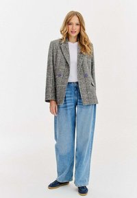 Mladá žena s dlouhými blond vlasy, oblečená v šedém kostkovaném saku, bílém topu, širokých modrých džínách a tmavě modrých teniskách, stojící na bílém pozadí.