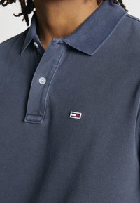 Polo in cotone blu scuro con finitura testurizzata, dotata di una patta con tre bottoni e un piccolo logo ricamato in rosso e bianco.