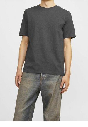 Man draagt een eenvoudige donkergrijze T-shirt met korte mouwen en vervaagde losse blauwe spijkerbroek, staat met ontspannen armen langs het lichaam.