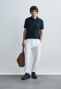 Navy polo shirt with short sleeves, white pleated pants, black sneakers, and a brown leather handbag. Smooth fabric and casual design.