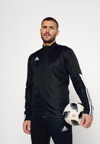 Zwarte sportjack met witte accenten, ritssluiting aan de voorkant, hoge kraag en lange mouwen, gecombineerd met een zwart-wit voetbal.