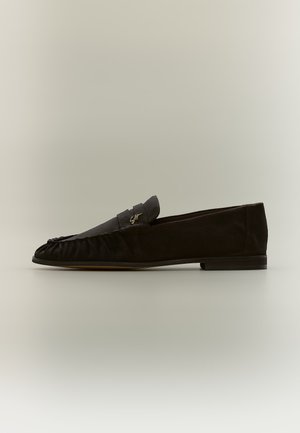 Mocassin plat en daim noir avec petit talon et petit détail logo métallique sur le côté, présenté en profil latéral sur fond neutre.