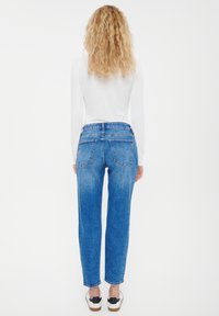 Kvinna med lockigt blont hår som bär en vit långärmad topp, blå jeans och vita sneakers, stående vänd mot en vit bakgrund.