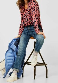 Blouse à imprimé léopard rose, jeans bleus avec revers, et baskets blanches. Une veste matelassée bleu clair est posée à côté d'elle sur un tabouret.
