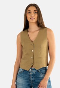 Chaleco de lino caqui con cuello en V, cinco botones y dos bolsillos frontales. Lleva encima de pantalones de mezclilla azul, que presentan un corte ajustado y un diseño elegante.