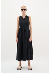 Schwarzes ärmelloses Kleid mit V-Ausschnitt und geraffter Taille, aus glattem Stoff gefertigt, mit einem fließenden Rock. Ergänzt durch beige Sandalen.