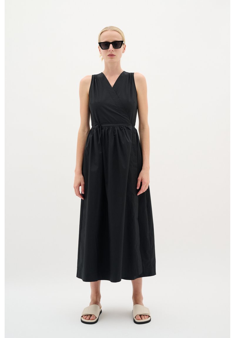 Schwarzes ärmelloses Kleid mit V-Ausschnitt und geraffter Taille, aus glattem Stoff gefertigt, mit einem fließenden Rock. Ergänzt durch beige Sandalen.