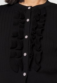 Černý cardigan vyrobený z žebrované pleteniny. Obsahuje centrální volánový detail a dekorativní stříbrné knoflíky se zirkony.