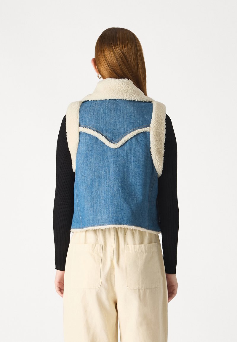 Jeansowa kamizelka z kremowym kołnierzem z sztucznego futra, z zaokrąglonymi szwami i surowym wykończeniem. Noszona na czarnej koszuli z długim rękawem i beżowych spodniach.