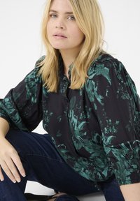 Camicia nera con motivo floreale verde, con bottoni davanti e maniche arricciate. Modello in jeans di denim, seduto su uno sfondo bianco.