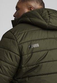 Giacca a vento verde oliva con motivo trapuntato, dotata di cappuccio e logo riflettente "PUMA" sulla spalla sinistra.