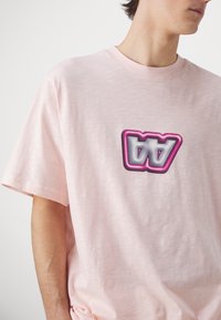 Camiseta de algodón de color rosa claro, con mangas cortas y cuello redondo, que presenta un logotipo multicolor prominente en el pecho.