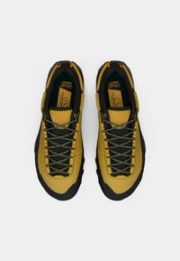 Par de botas de caminhada amarelas e pretas com atacadores verdes e laranjas, vistas de cima sobre um fundo branco.