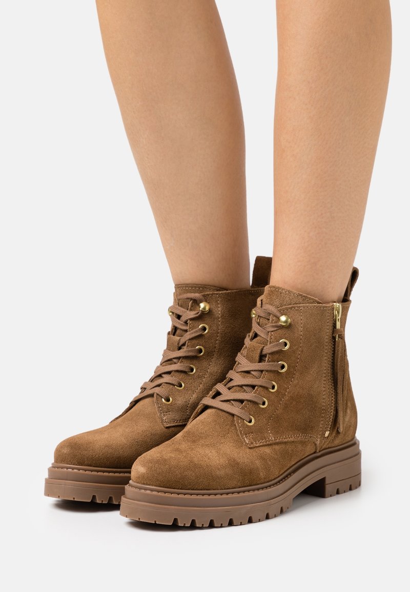 Ruske nubuk ankle boots dengan ujung bulat, hardware emas, tali di bagian depan, dan ritsleting samping. Sol karet tebal dengan pola tertentu.