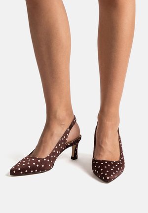 Tacones slingback marrones de punta fina con lunares blancos, usados sobre piernas desnudas contra un fondo liso.