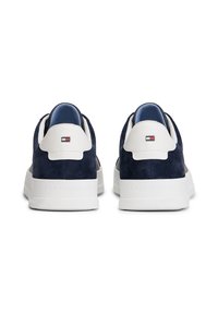 O pereche de sneakers din suede bleumarin cu tab-uri alb din piele la călcâi, cu branding embossat pe tălpile albe, văzuți din spate pe un fond alb.