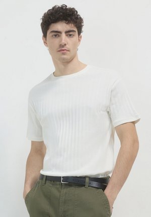 T-shirt bianca a righe con maniche corte e scollatura rotonda, indossata con pantaloni verdi e una cintura nera. Il tessuto ha un aspetto texturizzato.