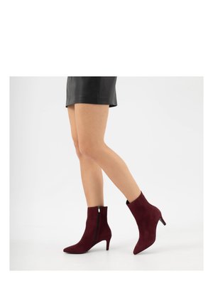 Frau trägt schwarzen Lederrock und burgunderrote Wildleder-Stiefeletten mit mittelhohen Absätzen und steht auf einem Bein vor weißem Hintergrund.