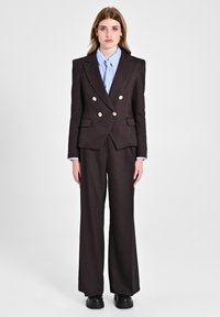 Blazer doppiopetto marrone sartoriale con bottoni dorati, abbinato a pantaloni a gamba larga coordinati e una camicia a righe azzurre sotto.