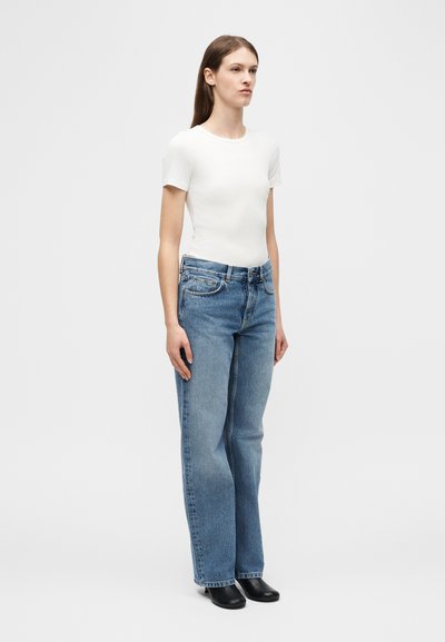 Νεαρή γυναίκα που στέκεται, φορώντας ένα απλό λευκό κοντομάνικο τοπ, ίσια μπλε τζιν με ψηλή μέση και μαύρα παπούτσια σε απλό ανοιχτό γκρι φόντο.