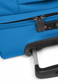 Blauer Rollkoffer mit strukturiertem Stoff, Doppelreißverschlüssen und ausziehbarem Griff mit Gummigriff. Schloss-System in der Nähe des Griffs sichtbar.