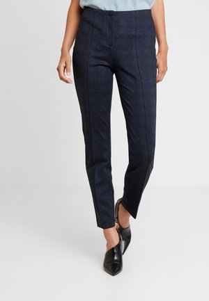 Kvinde iført mørkeblå slimfit bukser med frontsømme og sorte spidse slingback sko, stående foran en ensfarvet baggrund.