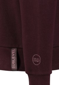 Burgundy sweatshirt with textured fabric, ribbed cuffs, and two labels: one textured and the other embossed with "SBLVL CNTMP."