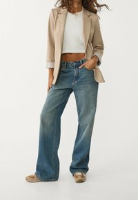 Blazer beige su top corto bianco, abbinato a jeans larghi blu sbiaditi e sneakers beige con dettagli bianchi.