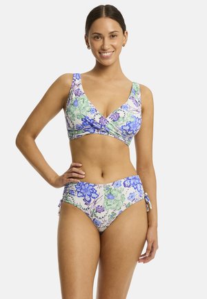 Lächelnde Frau in einem zweiteiligen Badeanzug mit blauen und grünen Blumen, steht mit einer Hand in der Hüfte vor weißem Hintergrund.