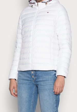 Femme portant une veste matelassée blanche à capuche avec fermeture éclair, petit logo sur la poitrine, et un jean taille haute bleu à braguette boutonnée.