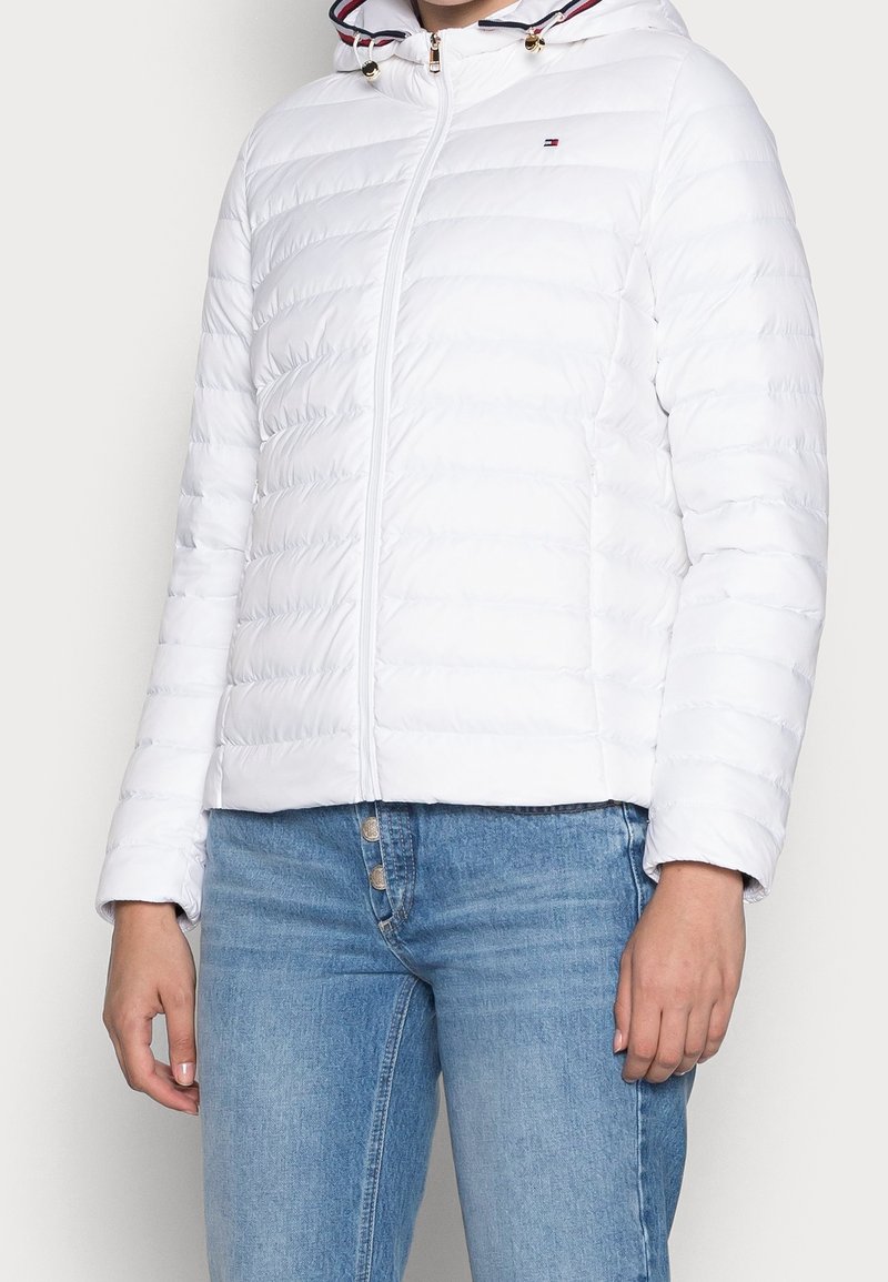 Femme portant une veste matelassée blanche à capuche avec fermeture éclair, petit logo sur la poitrine, et un jean taille haute bleu à braguette boutonnée.