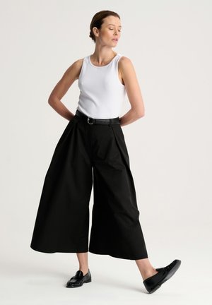 Kobieta w białym topie bez rękawów, czarnych szerokich spodniach skróconych, czarnym pasku i czarnych mokasynach stoi z jedną stopą wysuniętą do przodu.